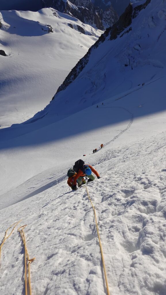 Ein Bergsteiger in leuchtender Ausrüstung erklimmt mithilfe von Seilen einen steilen, verschneiten Hang im Rahmen einer geführten Mont-Blanc-Expedition, während sich weiter unten am Berg weitere Teilnehmer befinden und im tiefen Schnee Spuren zu sehen sind.