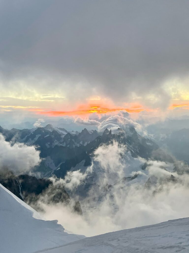 Die schneebedeckten Berggipfel, die an ein geführtes Abenteuer am Mont Blanc erinnern, sind bei Sonnenaufgang oder Sonnenuntergang in Wolken gehüllt, wobei ein orange-gelber Schimmer den Nebel durchbricht und eine dramatische und doch friedliche Landschaft entstehen lässt.