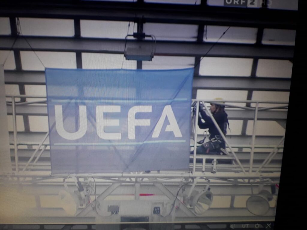 Ein Arbeiter in Schutzkleidung steht auf einem Gerüst hinter einem blauen Banner mit der Aufschrift „UEFA“ in weißen Buchstaben, das an einer Metallkonstruktion in einem Innenraum befestigt ist, und zeigt dabei Fachkenntnisse, die an Seilarbeiten im alpinen Gelände erinnern.