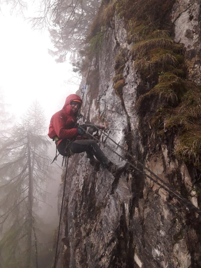 Eine Person in roter Jacke und mit Helm klettert in einem nebligen Wald eine steile, felsige Klippe hinauf und nutzt dabei Seile und Klettergurte zur Sicherheit – ein klassisches Beispiel für Seilarbeiten im alpinen Gelände.