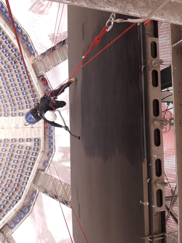 Ein Arbeiter mit Schutzhelm reinigt mithilfe von Seilen eine große Wand oder Konstruktion, während er hoch über dem Boden in einer Industrieanlage oder einem Stadion schwebt – ähnlich wie bei Seilarbeiten im alpinen Gelände, wo spezielle Seilzugangstechniken Sicherheit und Effizienz in extremen Höhen gewährleisten.
