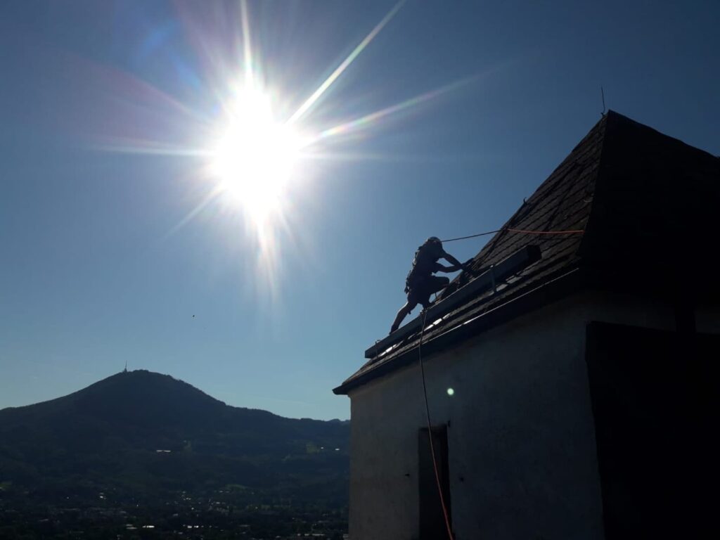 Eine Person ist als Silhouette zu sehen, während sie unter strahlendem Sonnenschein Seilarbeiten im alpinen Gelände auf einem steilen Dach ausführt, im Hintergrund sind Berge und ein klarer Himmel zu sehen.