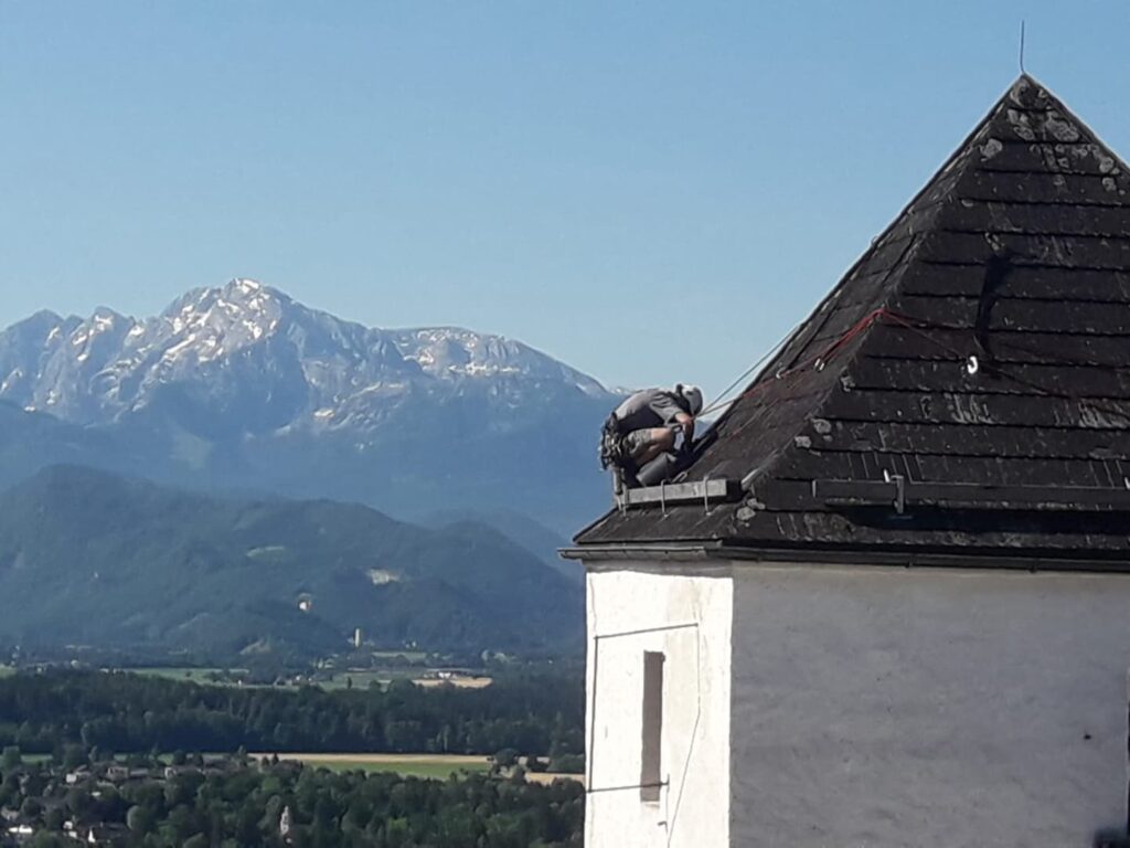 Ein Arbeiter in Schutzausrüstung führt Seilarbeiten im alpinen Gelände durch, während er das Dach eines weißen Gebäudes repariert oder inspiziert; im Hintergrund erstrecken sich majestätische Berge und grüne Täler unter einem strahlend blauen Himmel.