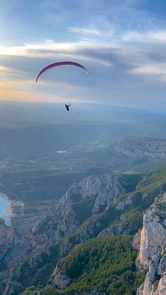 Ein „Hike & Fly“-Gleitschirmflieger schwebt bei Sonnenuntergang über schroffe Berge und einen sich schlängelnden blauen Fluss, vor dem sich eine weite Landschaft und dramatische Wolken abzeichnen.