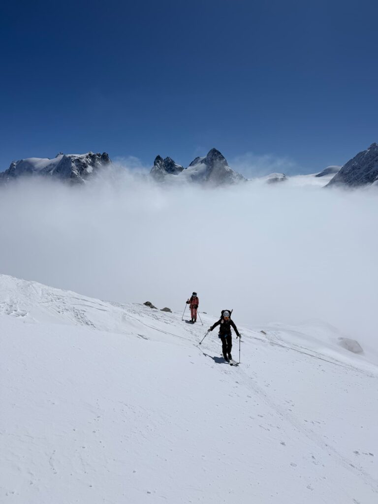 Zwei Personen mit Skistöcken wandern einen verschneiten Berghang hinauf und nehmen an geführten Skidurchquerungen in den Alpen teil, umgeben von dichtem Nebel und fernen, zerklüfteten Berggipfeln unter einem strahlend blauen Himmel.