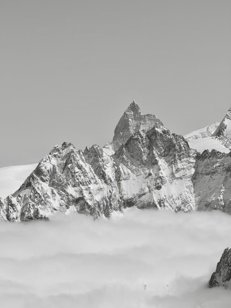 Schneebedeckte Berggipfel, darunter das berühmte Matterhorn, ragen unter einem klaren Himmel aus einer Wolkendecke empor und schaffen so eine dramatische und friedliche Landschaft in Schwarz-Weiß.