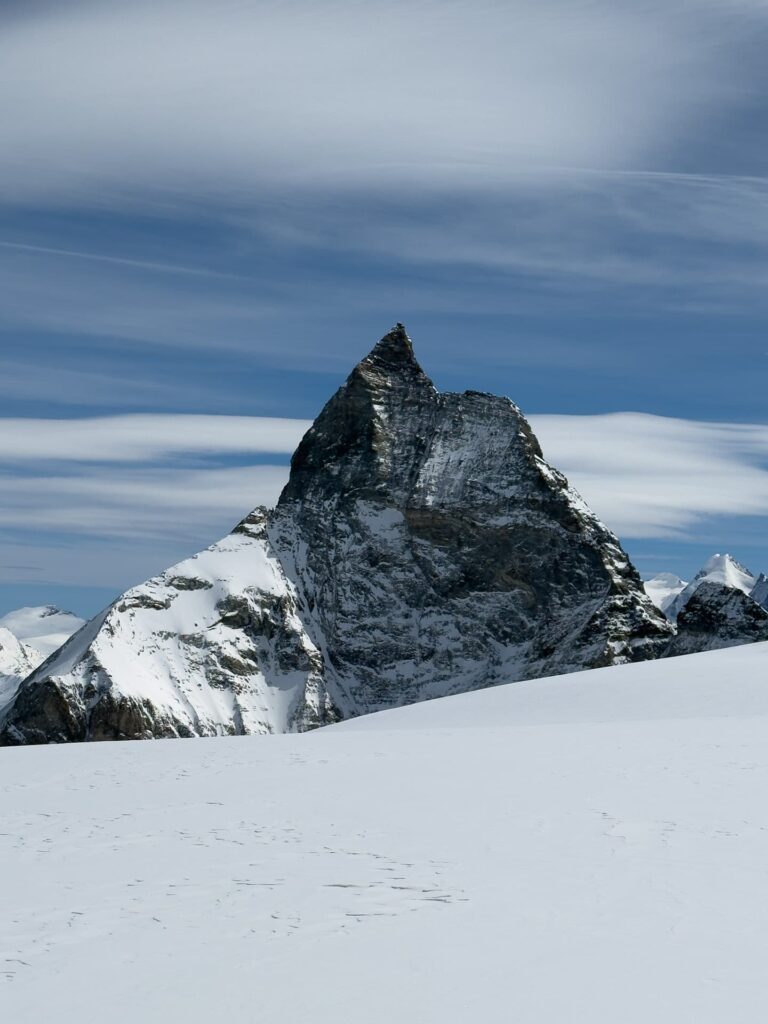 Ein hoher, zerklüfteter, schneebedeckter Berggipfel ragt vor einem blauen Himmel mit zarten Wolken empor und erinnert an eine Matterhorn-Expedition. Im Vordergrund bedeckt Schnee den Boden.
