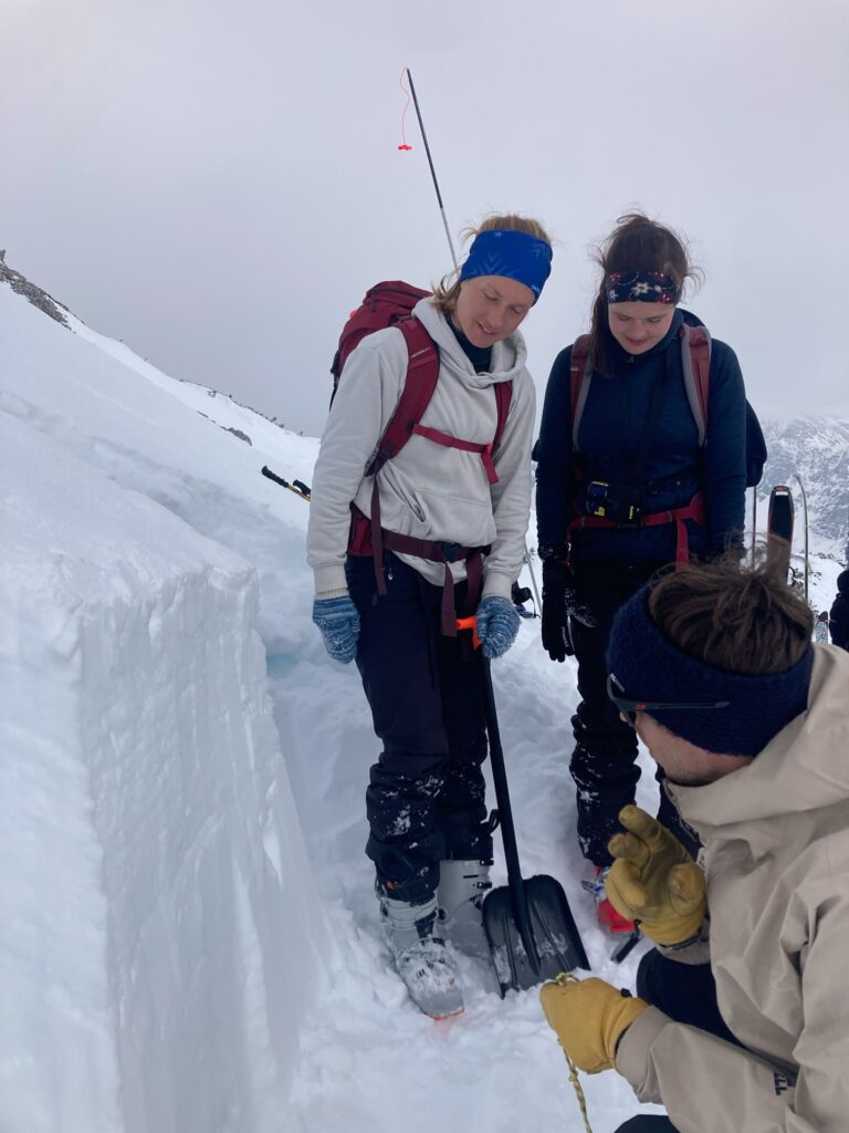 Drei Personen in Winterausrüstung untersuchen eine Schneegrube an einem verschneiten Berghang im Rahmen eines Lawinentrainings. Eine Person hält eine Schneeschaufel in der Hand, während eine andere kniet; möglicherweise besprechen sie während ihrer Wintertrainingssitzung die Schneeschichten oder die Lawinensicherheit.