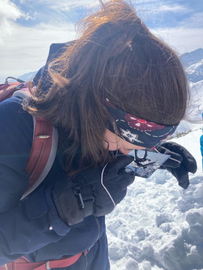 Eine Person, die ein Stirnband, Handschuhe und einen Wintermantel trägt, untersucht den Schnee mit einer Lupe, während sie im Rahmen des Lawinentrainings im Winter im Freien in einer verschneiten Berglandschaft steht.