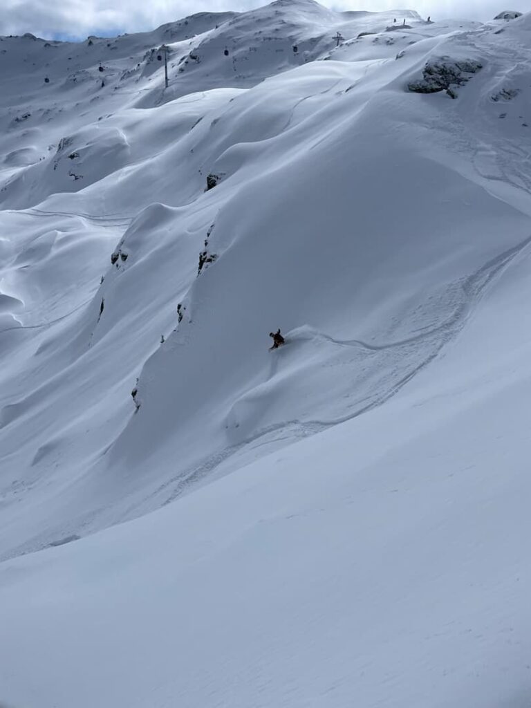 Ein Snowboarder gleitet unter bewölktem Himmel durch frischen, unberührten Schnee auf einem steilen, weitläufigen Berghang und verkörpert damit den Geist der „Freeride Touren Alpen“.