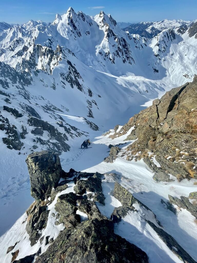 Ein Bergsteiger erklimmt einen verschneiten Berghang, umgeben von schroffen, schneebedeckten Gipfeln unter strahlend blauem Himmel. Im Vordergrund sind Felsvorsprünge zu sehen, die den Abenteuergeist geführter Skitouren in dieser atemberaubenden Alpenlandschaft einfangen.