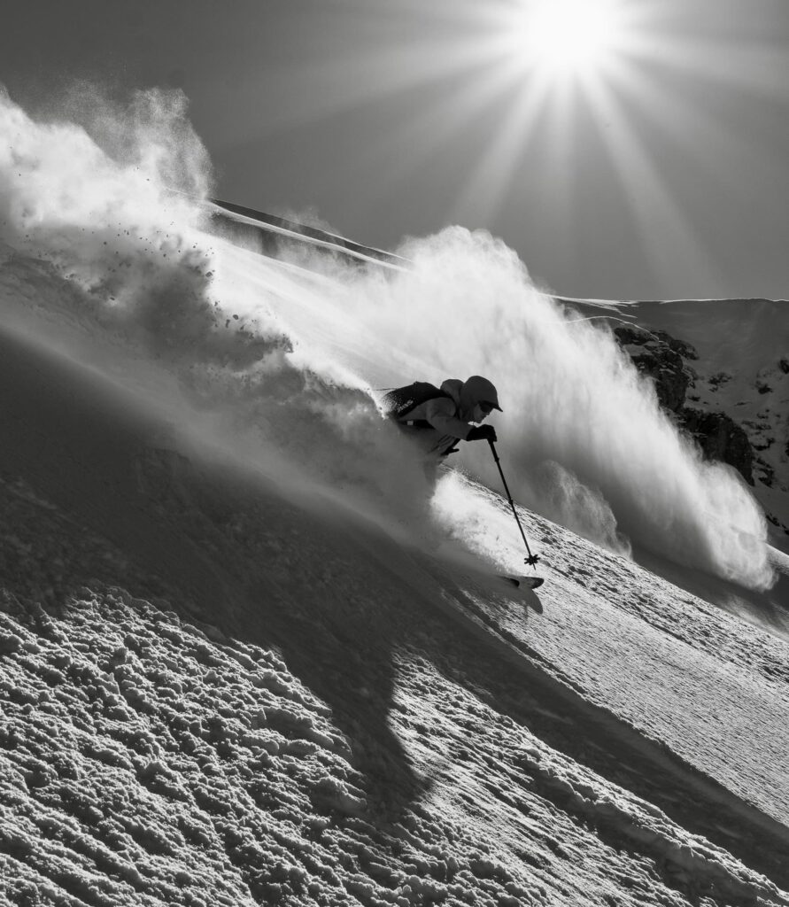 Ein Skifahrer saust einen steilen, verschneiten Hang auf einer der berühmten Freeride-Touren der Alpen hinunter und wirbelt dabei eine Wolke aus Pulverschnee auf, während die Sonne hoch am Himmel strahlt.