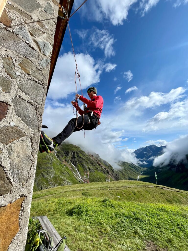 Eine Person in Kletterausrüstung seilt sich unter Anleitung von Alpincoaching Sommer an einer Steinwand ab, vor einer malerischen Kulisse aus Bergen, grünen Hügeln und einem teilweise bewölkten blauen Himmel.