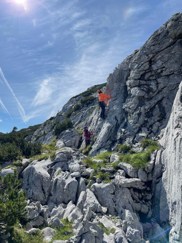 Zwei Menschen erklimmen einen felsigen Berghang; einer von ihnen befindet sich weiter oben und trägt eine orangefarbene Jacke. Dieser anspruchsvolle Aufstieg könnte Teil eines „Alpincoaching Sommer“-Programms sein. Über ihnen ist ein strahlend blauer Himmel mit Kondensstreifen zu sehen. Zwischen den Felsen wachsen Grasflecken und kleine Sträucher.