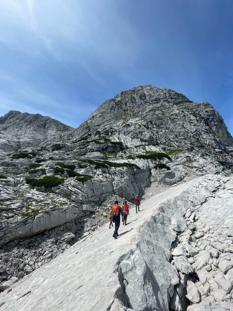 Drei Wanderer mit Rucksäcken steigen unter strahlend blauem Himmel einen felsigen Bergpfad hinauf, umgeben von großen grauen Felsformationen und spärlicher Vegetation – eine ideale Kulisse für die Abenteuer von Alpincoaching Sommer.