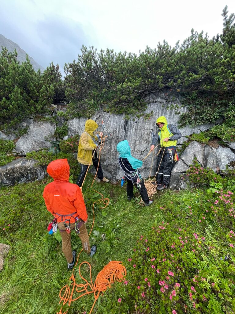 Vier Personen in bunten Jacken stehen auf einer Wiese in der Nähe von Felsen, halten Kletterseile und Ausrüstung in den Händen und scheinen sich während einer „Alpincoaching Sommer“-Veranstaltung auf eine Klettertour im Grünen vorzubereiten oder diese zu besprechen.