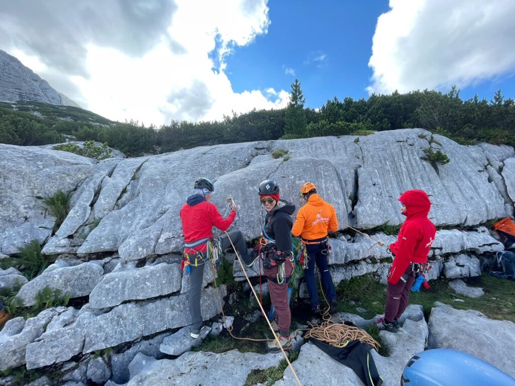 Vier Kletterer mit Helmen und Outdoor-Ausrüstung bereiten ihre Ausrüstung am Fuße einer felsigen Klippe vor, umgeben von Grün und unter einem teilweise bewölkten Himmel. Während dieser „Alpincoaching Sommer“-Veranstaltung liegen ein blauer Helm und Seile in der Nähe auf dem Boden, bereit für ihr nächstes Abenteuer.