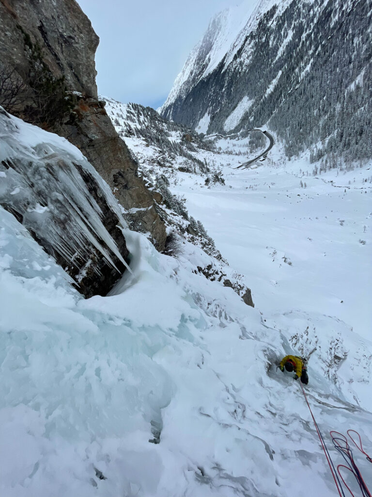 Ein Kletterer in gelber Kleidung übt sich im Eisklettern, während er mit Seilen einen vereisten, schneebedeckten Berghang mit gefrorenen Wasserfällen erklimmt. Im Hintergrund sind eine kurvenreiche Straße und ein verschneites Tal zu sehen.