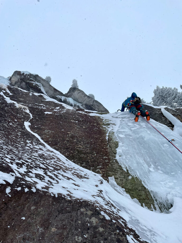 Ein Kletterer in Winterausrüstung erklimmt mithilfe von Seilen und Steigeisen eine steile, vereiste Felswand, umgeben von Schnee und bewölktem Himmel – ein Bild, das die enorme Herausforderung des Eiskletterns unter rauen alpinen Bedingungen verdeutlicht.