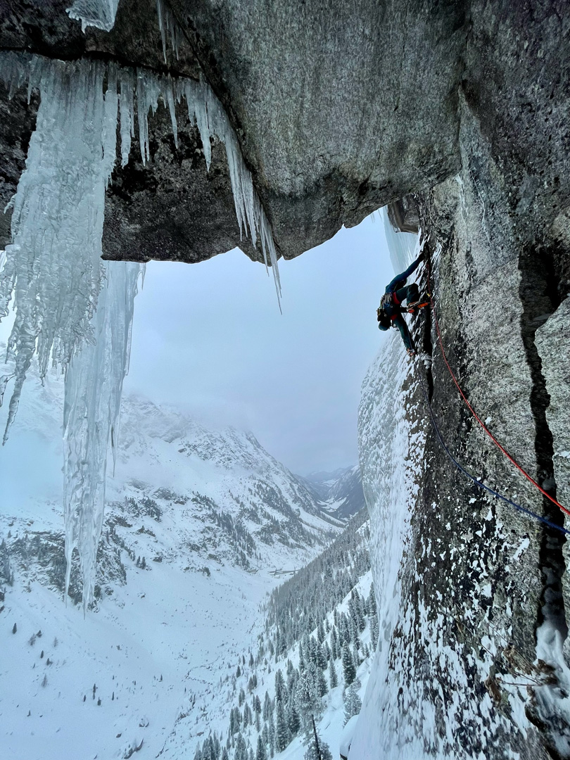 Ein Kletterer betreibt Eisklettern, während er eine steile, vereiste Felswand mit großen Eiszapfen darüber erklimmt, eingebettet in eine verschneite Berglandschaft. Gesichert durch ein rotes Seil, ist er umgeben von winterlicher Kulisse und weit entfernten, schneebedeckten Bäumen.