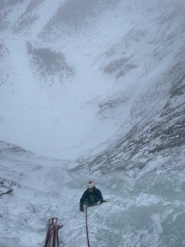 Ein Kletterer erklimmt einen steilen, vereisten Berghang bei Schnee und Nebel, hält sich an Seilen fest und ist dabei von schneebedeckten Felsen und einer nebligen Winterlandschaft umgeben – ein eindrucksvoller Beweis für die Herausforderungen des Eiskletterns.