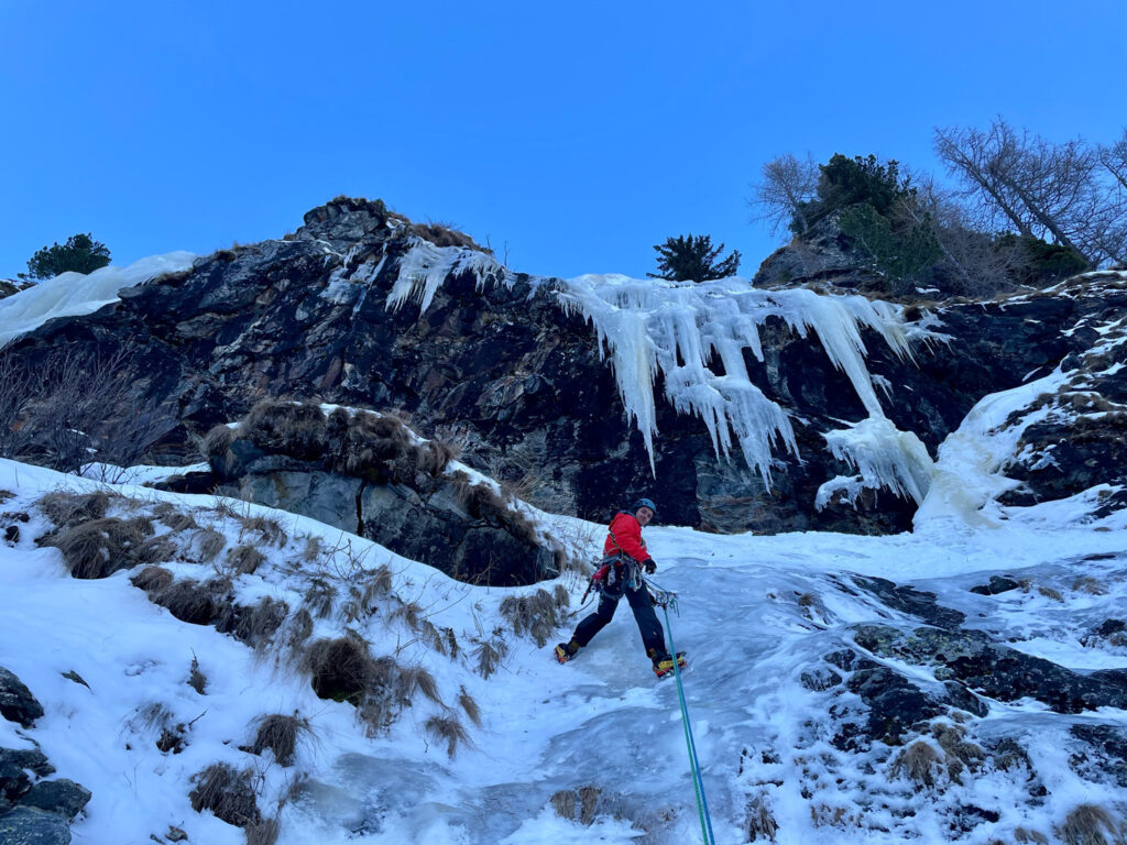 Eine Person in roter Kleidung betreibt Eisklettern, während sie unter strahlend blauem Himmel einen verschneiten, vereisten Hang erklimmt, an dessen Felswänden Eiszapfen herabhängen.