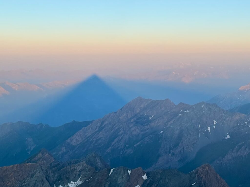 Berggipfel bei Sonnenaufgang, deren dreieckiger Schatten auf die nebelverhangenen Berge in der Ferne fällt und sich so als markante blaue Silhouette vor dem orange-rosa Himmel abhebt. Auf dem schroffen, felsigen Gelände sind einige Schneeflecken zu erkennen, die an ein klassisches alpines Abenteuer auf einem 3000er im Salzburger Land erinnern.