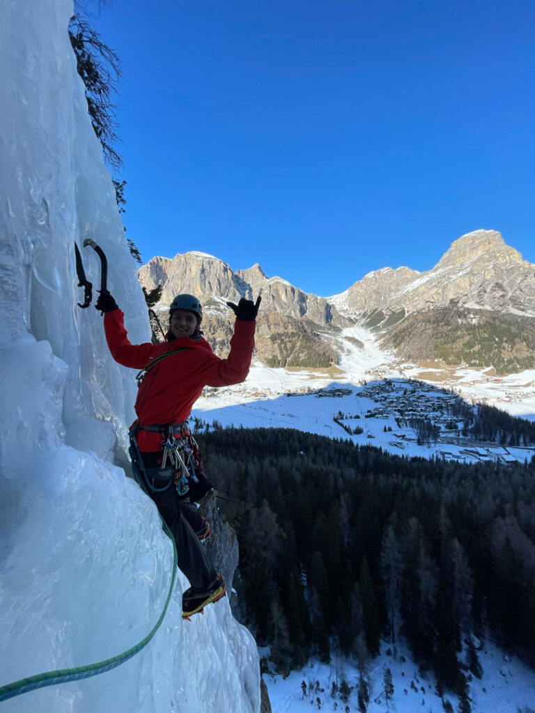 Ein Kletterer in roter Ausrüstung erklimmt mit Eispickeln eine vereiste Felswand, lächelt dabei und zeigt das Peace-Zeichen – ein perfekter Moment, der den Nervenkitzel des Eiskletterns einfängt. Im Hintergrund erstrecken sich unter einem strahlend blauen Himmel schneebedeckte Berge und ein bewaldetes Tal.