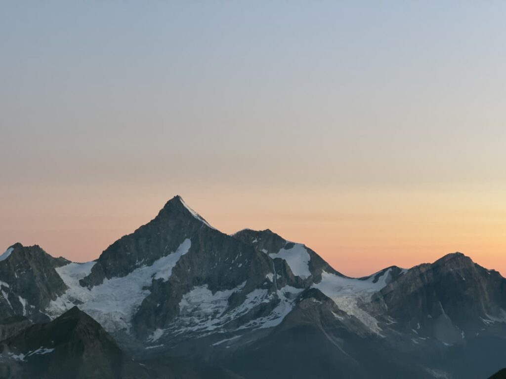 Schneebedeckte Berggipfel unter klarem Himmel bei Sonnenuntergang, mit sanften Orange- und Rosatönen am Horizont – ein unvergesslicher Anblick, der an eine geführte Besteigung des Weisshorns erinnert.