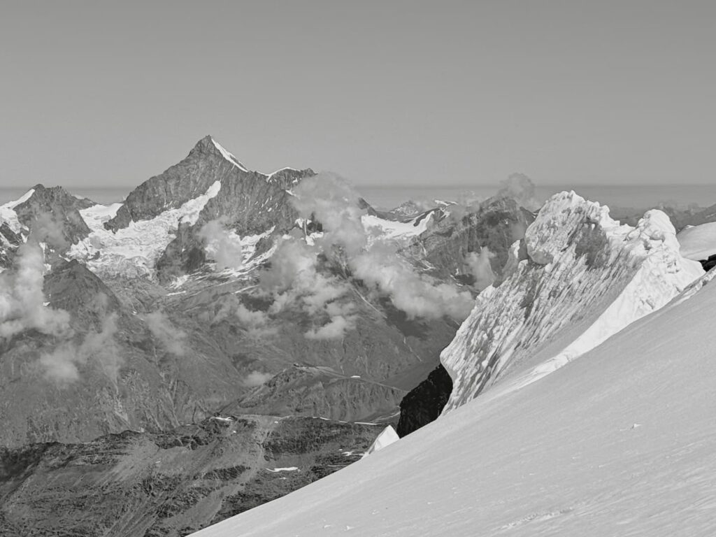 Schwarz-Weiß-Foto von schneebedeckten Berggipfeln. Im Vordergrund ist ein verschneiter Hang zu sehen, während sich im Hintergrund schroffe Berge – darunter das Weisshorn – dramatisch erheben, während Wolken unter einem klaren Himmel dahinziehen.