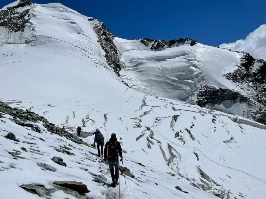 Drei Menschen wandern unter strahlend blauem Himmel einen verschneiten, felsigen Berghang hinauf, umgeben von Gletschern und schroffen Gipfeln auf der berühmten Spaghetti-Tour am Monte Rosa.