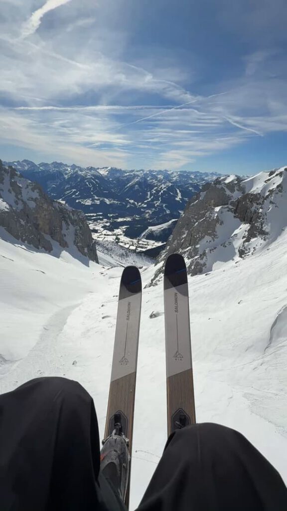 Aus der Perspektive eines Skifahrers sieht man zwei Skier auf einem verschneiten Berghang, im Hintergrund rauer Alpengipfel und ein Tal unter blauem Himmel – eine ideale Kulisse für ein unvergessliches „Hike & Fly“-Abenteuer mit Führung.