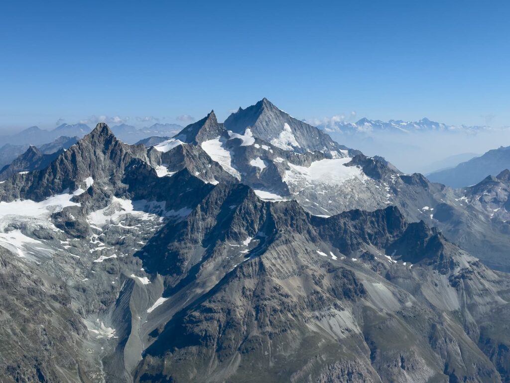 Luftaufnahme von schroffen, felsigen Bergen mit Schneeflecken und Gletschern unter einem strahlend blauen Himmel, wobei entfernte Gipfel – wie jene, die man bei einer geführten Besteigung des Weisshorns zu sehen bekommt – durch einen leichten Dunst hindurch sichtbar sind.