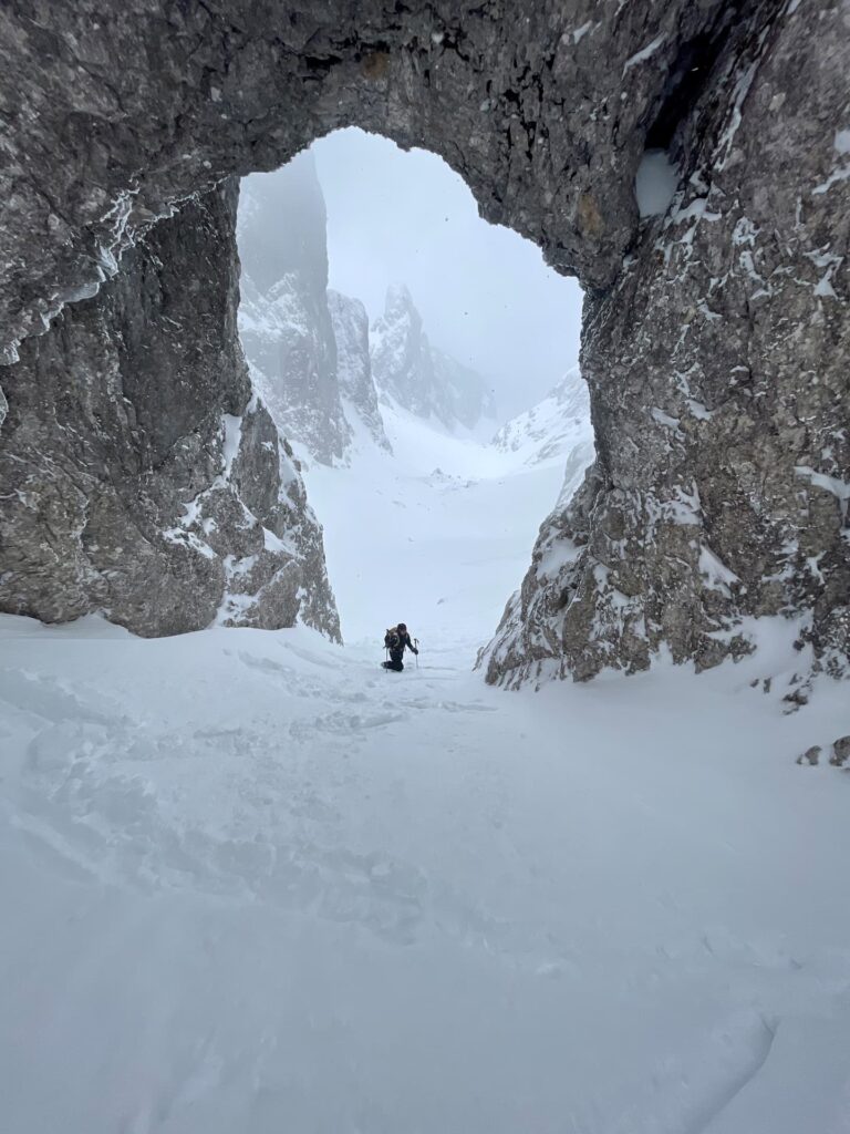 Ein Bergsteiger erklimmt einen verschneiten, steilen Berghang, der von einem großen natürlichen Felsbogen eingerahmt wird, während im Hintergrund schneebedeckte Gipfel und ein bewölkter Himmel zu sehen sind – eine atemberaubende Szene, die an geführte Skidurchquerungen in den Alpen erinnert.