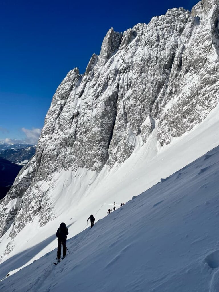 Mehrere Bergsteiger erklimmen unter strahlend blauem Himmel einen steilen, verschneiten Berghang, während sich neben ihnen schroffe, schneebedeckte Felsen steil emporragen – eine inspirierende Szenerie, wie man sie oft auf geführten Skitouren antrifft.