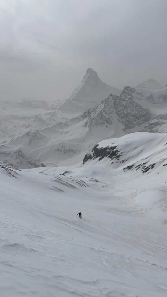 Ein Skifahrer fährt unter bewölktem Himmel einen verschneiten Berghang hinunter, im Hintergrund sind schroffe, schneebedeckte Gipfel und ein markanter Berg zu sehen – eine Szene, die an geführte Skihochtouren in den Alpen erinnert.