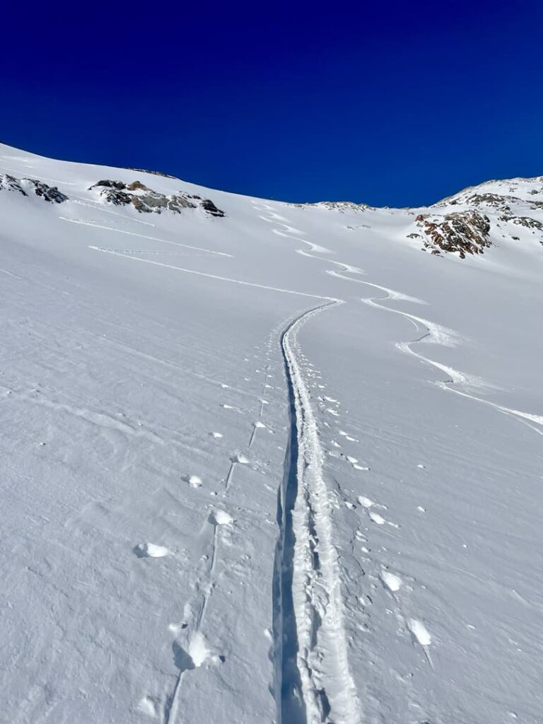 Eine einzelne Skispur schlängelt sich unter einem klaren, tiefblauen Himmel einen verschneiten Berghang hinauf; im Schnee sind vereinzelte Felsen und weitere, kaum erkennbare Skispuren zu sehen – eine einladende Szene, die das Wesen geführter Skitouren in unberührten Alpenlandschaften einfängt.