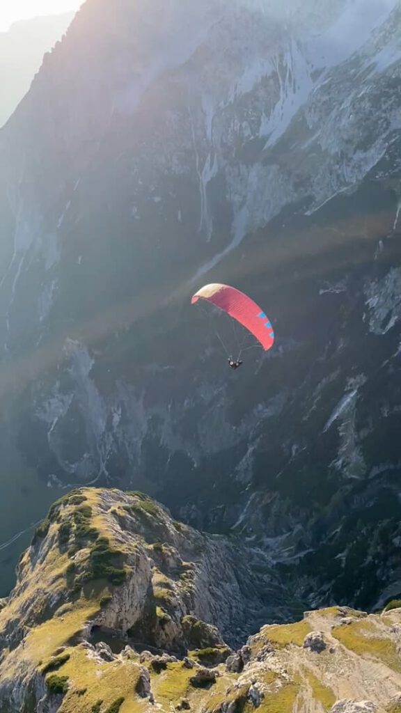 Eine Person gleitet mit einem rot-blauen Gleitschirm bei sanftem Sonnenlicht über steile, felsige Berge und grüne Bergrücken und genießt ein unvergessliches „Hike & Fly“-Abenteuer.