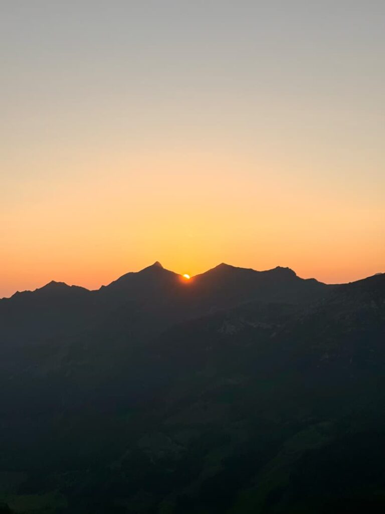 Die Sonne geht hinter einer Kette dunkler Berge unter – vielleicht sind es einige der mächtigen 3000er des Salzburger Landes –, und taucht den Himmel in ein warmes Orange, während der Vordergrund im Schatten bleibt.