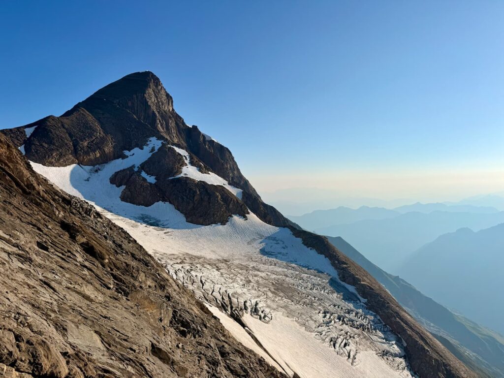Ein schroffer Berggipfel mit Schnee- und Eisflecken ragt majestätisch empor und bietet atemberaubende Ausblicke, wie sie für eine geführte Tour auf einen 3000er im Salzburger Land typisch sind: In der Ferne zeichnen sich unter einem strahlend blauen Himmel verschwommene Bergkonturen ab.