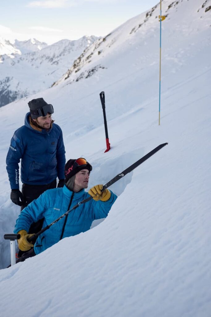 Zwei Personen in Winterausrüstung untersuchen während einer Lawinen-Wintertrainingsveranstaltung mit Hilfe von Werkzeugen die Schneeschichten in einem Graben an einem verschneiten Berghang. Eine Person kniet und hält eine Säge in der Hand, während die andere hinter ihr steht. Im Hintergrund sind Skier und Berge zu sehen.