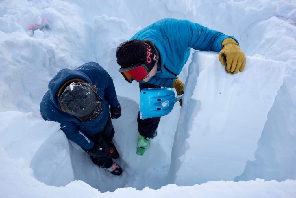 Zwei Personen in Winterkleidung stehen in einem tiefen Schneegraben; eine von ihnen hält eine blaue Schaufel in der Hand und untersucht einen großen Schneeblock. Sie sind von Schnee umgeben und scheinen im Rahmen einer Lawinen-Winterausbildung eine Analyse oder Ausgrabung durchzuführen.