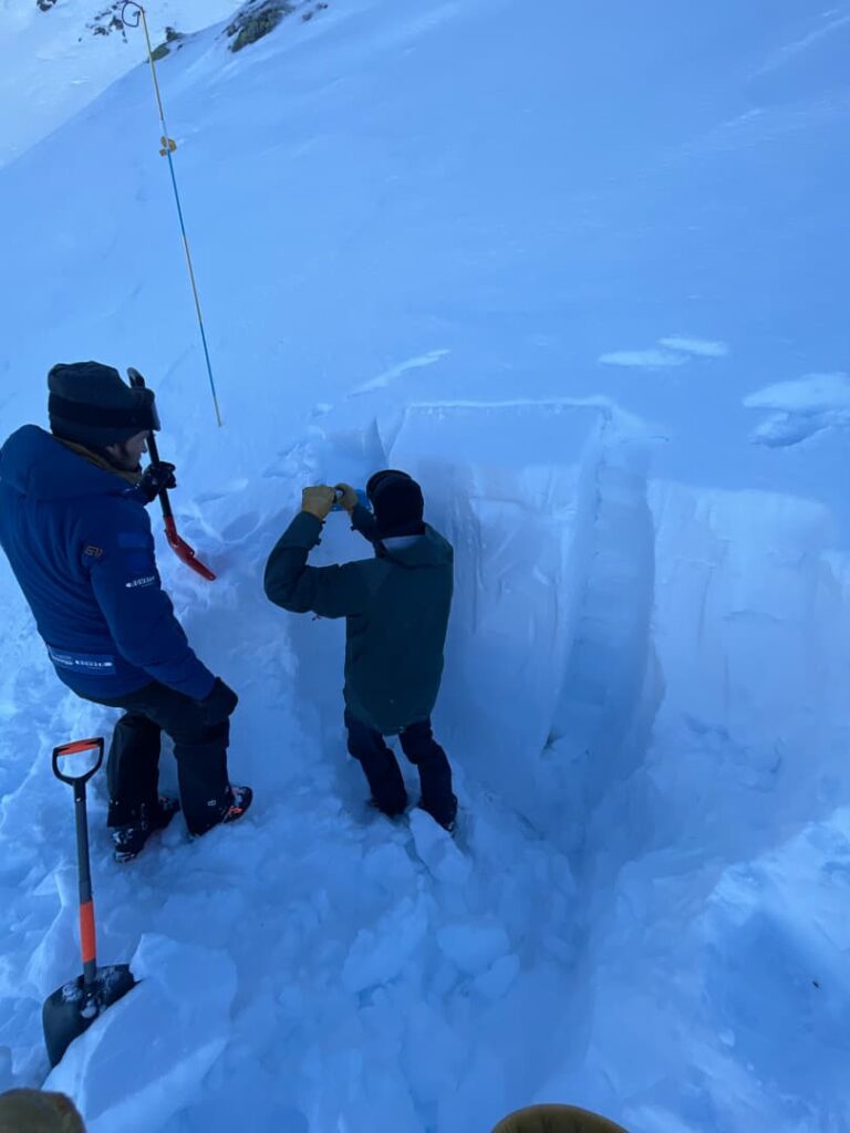 Zwei Personen in Winterkleidung untersuchen eine tiefe Schneegrube an einem Hang und analysieren vermutlich die Schneeschichten. Es sind Schaufeln und Ausrüstung zu sehen, was auf eine Lawinenausbildung hindeutet – Winteraktivitäten mit Schwerpunkt auf der Lawinengefahrenbeurteilung.