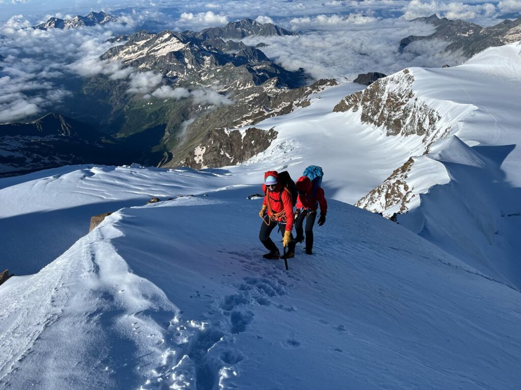 Zwei Bergsteiger in roten Jacken erklimmen einen verschneiten Bergrücken, umgeben von schneebedeckten Gipfeln und Wolken unter ihnen, unter einem klaren Himmel – eine inspirierende Szene, die an eine Lyskamm-Expedition erinnert.