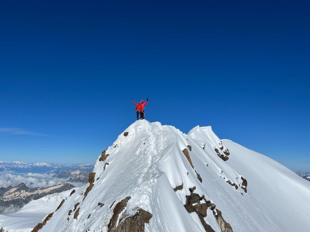 Eine Person in einer roten Jacke steht triumphierend auf einem verschneiten Berggipfel unter strahlend blauem Himmel und hält ihre Trekkingstöcke hoch – sie feiert den erfolgreichen Aufstieg auf den Lyskamm mit Führung.