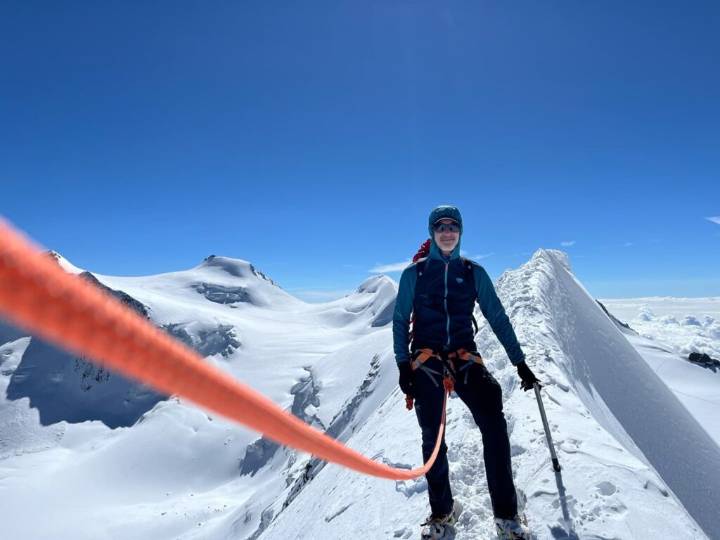 Ein Bergsteiger in Winterausrüstung steht während einer geführten Lyskamm-Besteigung auf einem verschneiten Berggrat, hält einen Eispickel in der Hand und ist unter strahlend blauem Himmel mit einem orangefarbenen Seil gesichert. Schneebedeckte Gipfel erstrecken sich bis in die Ferne.