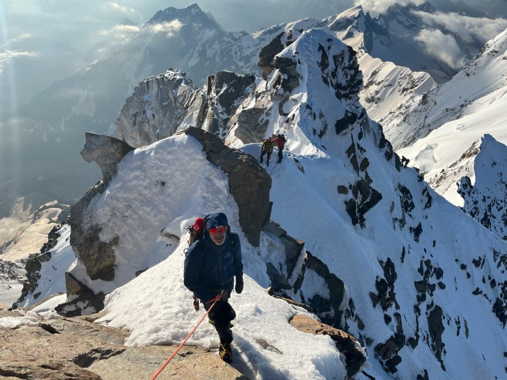 Drei Bergsteiger in Winterausrüstung erklimmen im Rahmen einer geführten Expedition zur Dom- und Dufourspitze einen schmalen, schneebedeckten Bergrücken mit Felsvorsprüngen, umgeben von imposanten Gipfeln und Wolken.