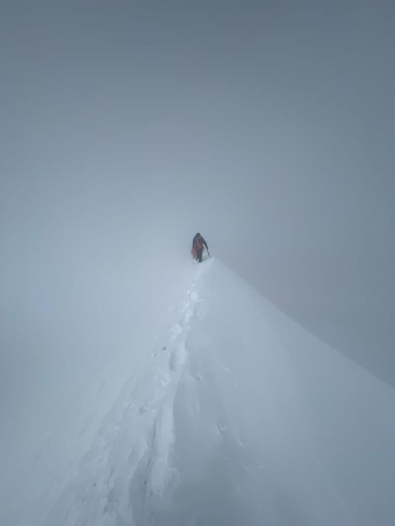Ein einzelner Bergsteiger in roter Ausrüstung wandert auf einem schmalen, verschneiten Grat, umgeben von dichtem Nebel, und hinterlässt Fußspuren auf dem schneebedeckten Weg – eine Szene, die an eine geführte Besteigung des Lyskamm durch die hochalpine Wildnis erinnert.