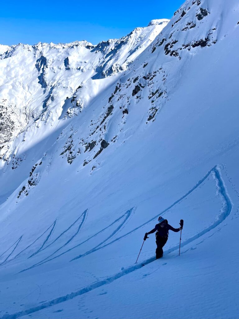 Ein Skifahrer steigt einen verschneiten Berghang hinauf und hinterlässt dabei geschwungene Spuren – ein Abenteuer, das an geführte Skihochtouren in den Alpen erinnert. Im Hintergrund ragen schroffe, schneebedeckte Gipfel unter einem strahlend blauen Himmel empor.