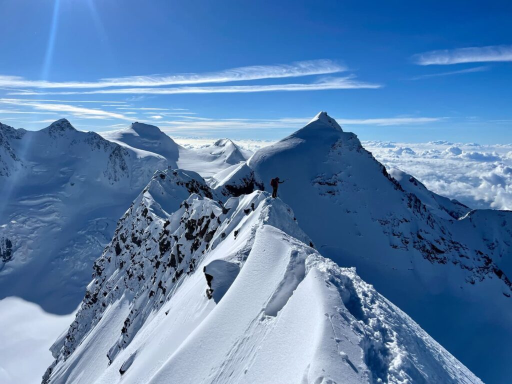Eine Person steht auf einem verschneiten Bergrücken, umgeben von hoch aufragenden, schneebedeckten Gipfeln unter einem klaren blauen Himmel mit vereinzelten Wolken, und erlebt den Nervenkitzel eines Lyskamm-Abenteuers.